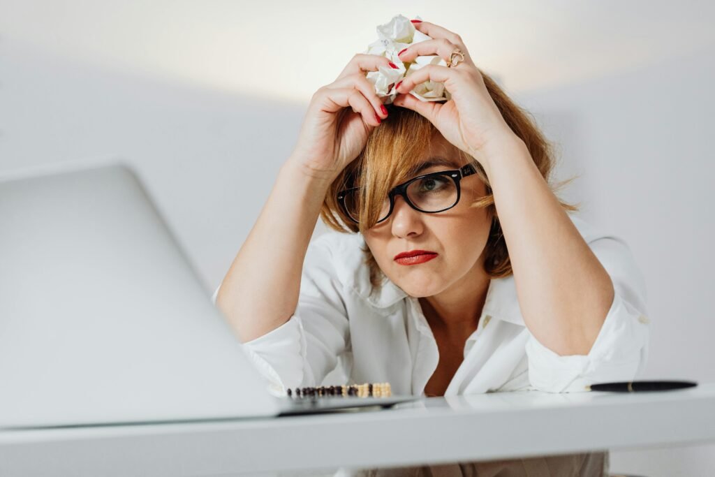 Flex Printing Services In Dubai | Flex Printing Near Me Frustrated woman with eyeglasses at a desk, crumpling paper while using a laptop indoors.